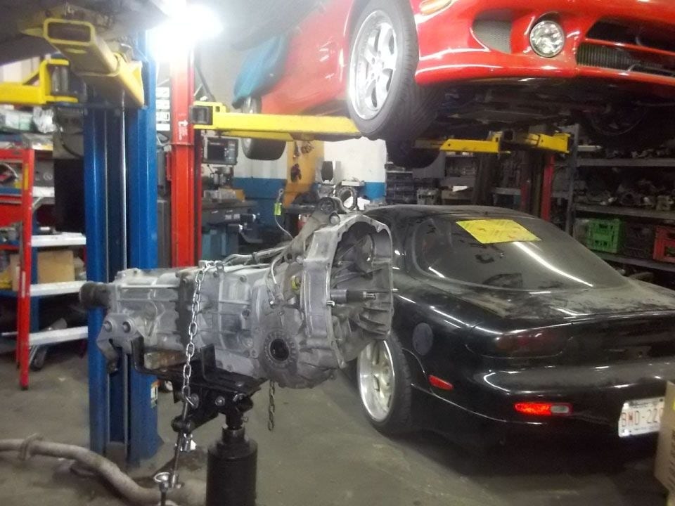 The Clutch-Pro shop floor: a transmission lowered on a jack under a black RX-7, a red car up on a lift above